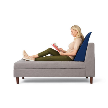 A woman reclines on a grey chaise lounge, reading a magazine with the Medline High-Density Foam Bed Wedge (24 x 20 x 8.5) in dark blue supporting her back, against a plain white background. She wears a beige top, green pants, and light shoes.