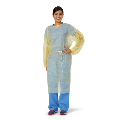 A person wearing a yellow Medline Disposable Full Back Spunbond Cover Gown over blue scrubs and black shoes stands smiling with one hand on their hip against a white background.