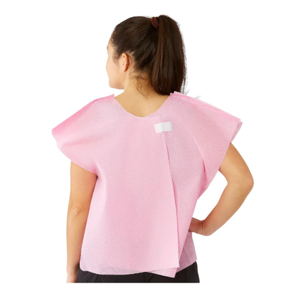 A woman with dark hair in a ponytail is seen from the back wearing a sleeveless pink Medline Disposable Multilayer Patient Cape (30" x 21"), fastened near the collar with a white adhesive strip.