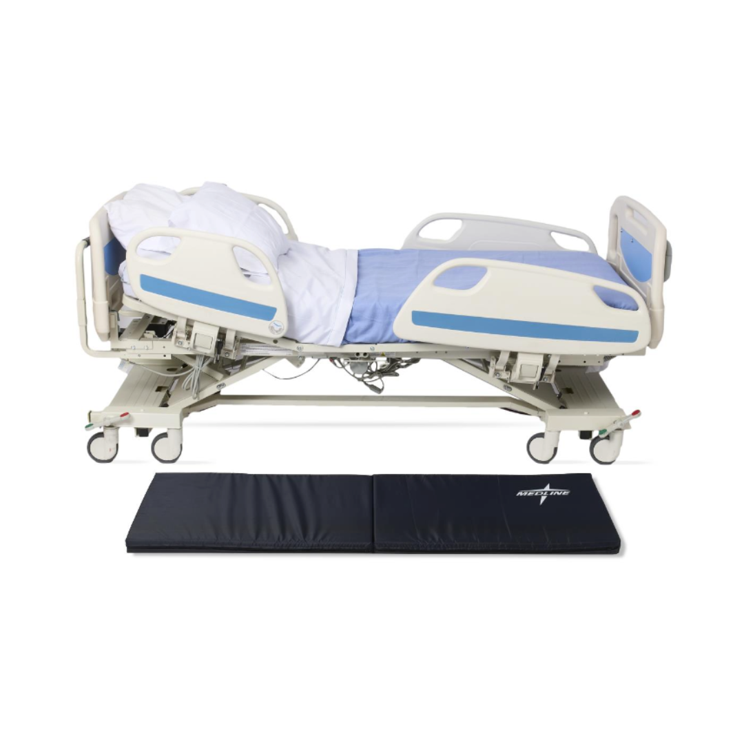 A hospital bed with blue and white bedding is reclined, while a Medline Bi-Fold Fall Mat is placed on the floor in front of it to provide effective fall protection.