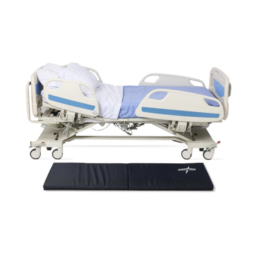 A hospital bed with blue and white bedding is reclined, while a Medline Bi-Fold Fall Mat is placed on the floor in front of it to provide effective fall protection.