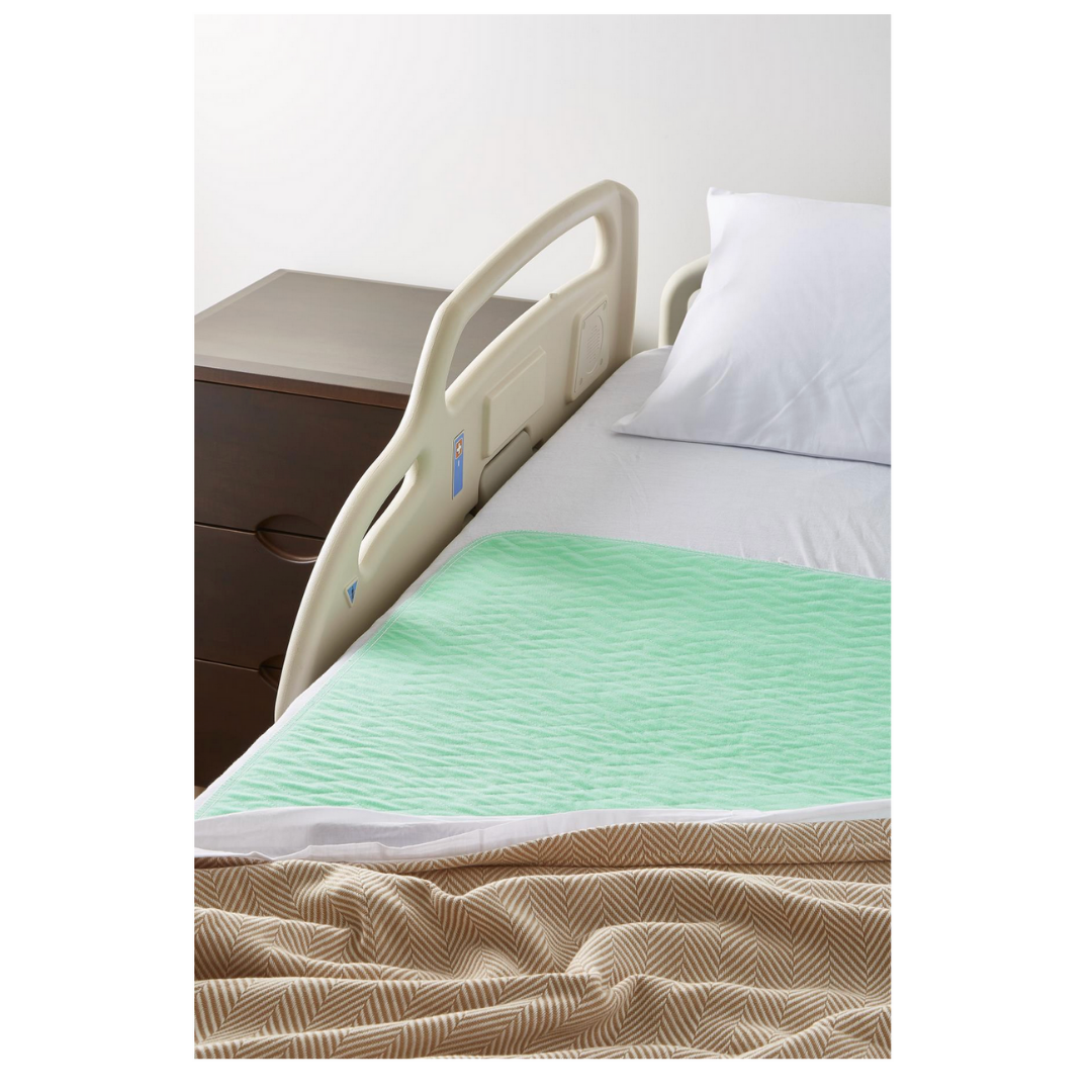 A hospital bed features white sheets, a white pillow, and a Medline Quick Dry Poly Laminated Reusable Underpad (34 x 36) with a green quick-dry surface. A brown nightstand sits nearby, and the bed has a light beige safety rail and patterned blanket.