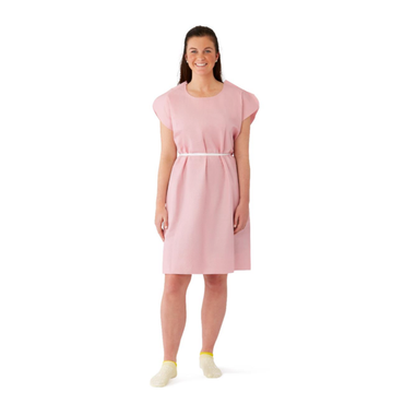 A smiling woman faces forward, wearing a light pink Medline Reinforced Tissue Patient Gown with a white belt and light yellow socks against a plain white background.