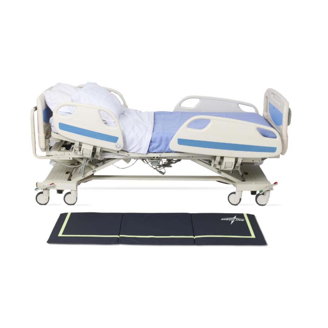 A hospital bed with side rails is in a semi-reclined position, covered with a blue and white blanket. Next to it on the floor is a Medline Trifold Beveled Edge Fall Mat with Glow featuring green edges.