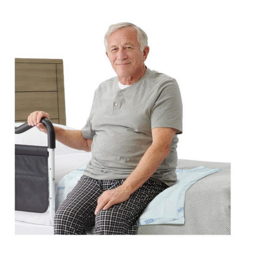 An older man in a gray shirt and plaid pajama pants sits on the bed edge, using a bed rail, with a Medline Ultrasorbs Advanced Premium Underpad (30 x 36) beneath him for effective incontinence management.