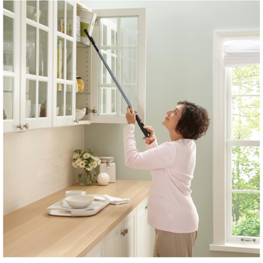 A woman uses the Medline 31" Reacher Grabber Tool with an ergonomic grip to pick an item from the top shelf of her glass-front kitchen cabinet while standing safely on the floor in a bright kitchen with flowers by the window.
