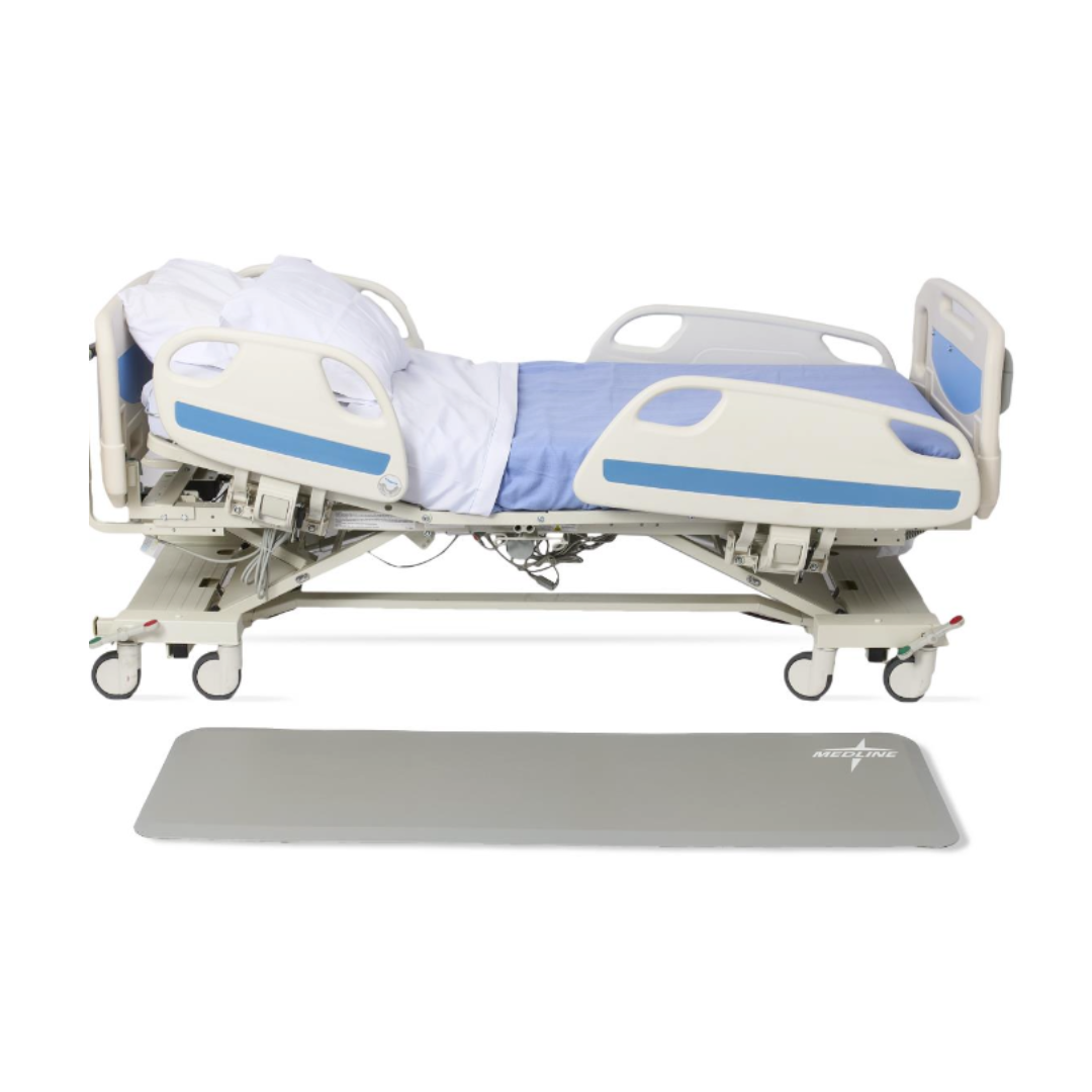 A hospital bed with side rails and a blue mattress is partially elevated, featuring white bedding and pillows; a Medline Beveled Edge Bedside Fall Mat is placed on the floor beside the bed for added safety.