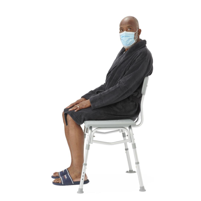 An older man wearing a face mask, dark robe, and blue slides sits upright on the Medline Bariatric Transfer Bath Bench with Back, featuring a blow-molded seat, against a plain white background.