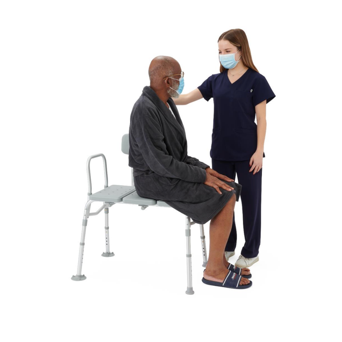 A healthcare worker in navy scrubs and a face mask stands beside an older man seated on a Medline Bariatric Transfer Bath Bench with Back. The worker rests a supportive hand on his shoulder as the man sits securely on the sturdy shower chair.