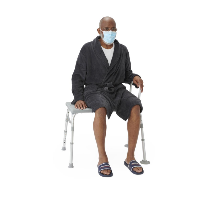 An older man in a face mask, glasses, slippers, and dark robe sits on the Medline Bariatric Transfer Bath Bench with Back—featuring a rust-resistant aluminum frame and blow-molded seat—against a plain white background.