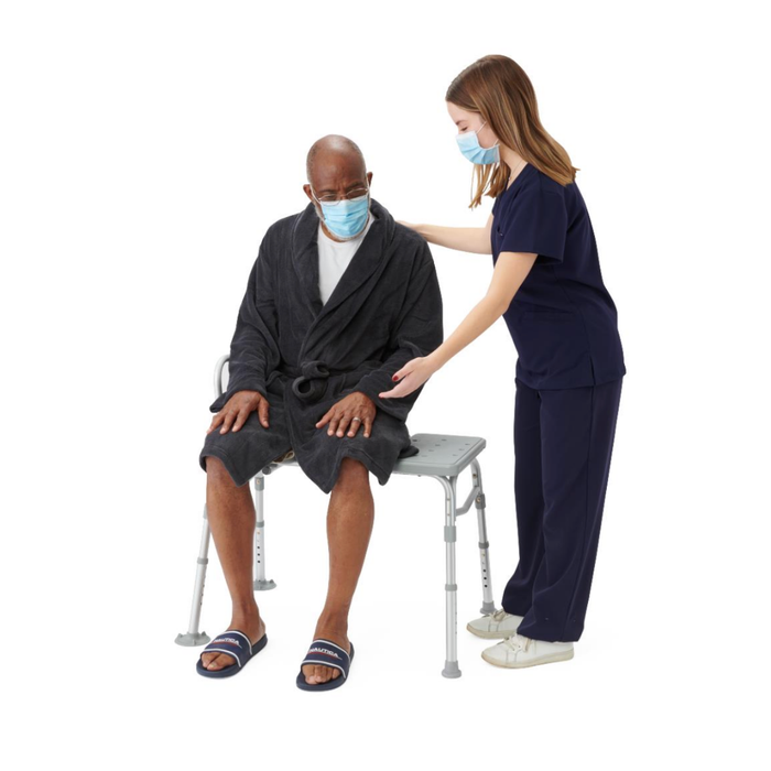 A young female healthcare worker in navy scrubs assists an older man in a bathrobe and slippers as he sits on a Medline Bariatric Transfer Bath Bench with Back; both wear face masks against a plain white background.