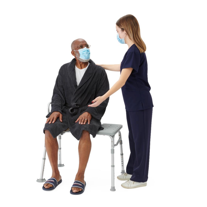 A healthcare worker in navy scrubs and a mask assists an older man in a robe, slippers, and mask, seated on a Medline Bariatric Transfer Bath Bench with Back featuring a blow-molded seat and rust-resistant aluminum frame against a white background.