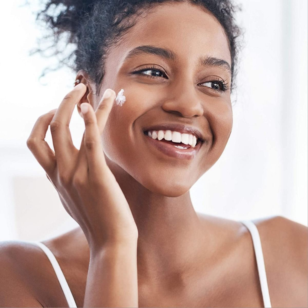 A smiling woman with curly hair applies Medline Remedy Clinical Skin Cream - Full Body Lotion to her cheek, wearing a white tank top in a bright, softly lit room.