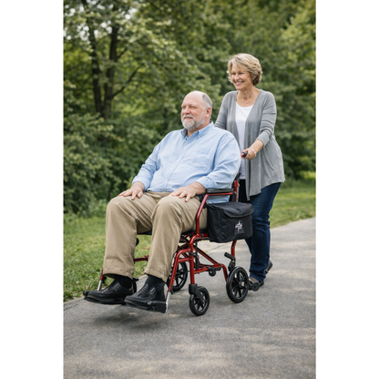 A smiling woman pushes an older man in a red Medline Steel Transport Chair with Wide 19" Seat along a paved park path surrounded by green trees.