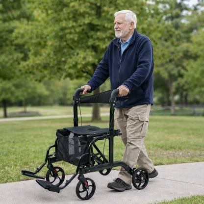 An older man with gray hair and a beard walks outdoors on a sidewalk using a Medline Euro Translator Hybrid Transport Chair and Rollator. He wears a navy jacket, beige pants, and brown shoes, with green grass and trees behind him.