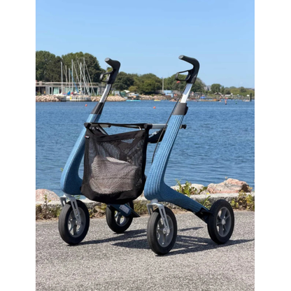 A Mesh Bag For ByAcre Rollators is attached to a modern blue rollator walker, which is parked on a paved path by a lake, surrounded by trees and rocks under a clear blue sky.