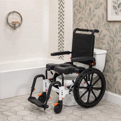 A MetaPal® M1-SP Self-Propelled Shower Commode Chair with large rear wheels, a black seat, and a backrest, positioned in a bathroom next to a toilet and bathtub.