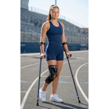 A young woman in athletic wear stands on a running track using the Millennial Medical in-Motion Pro Forearm Crutch Set and a black knee brace on her right leg, looking forward confidently with stadium bleachers behind her.