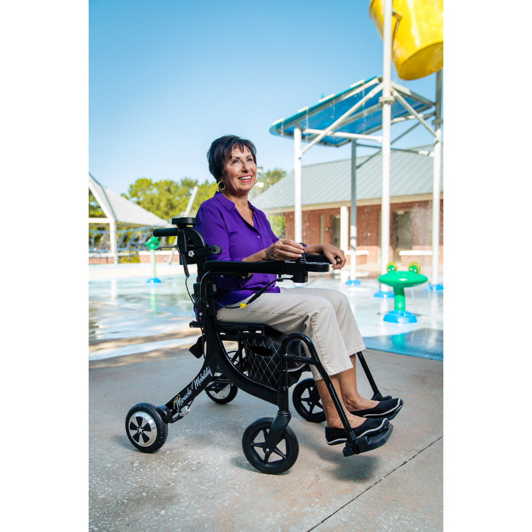 An older woman in a purple shirt and beige pants smiles while seated in a Miracle Mobility Portable 4N1 Walker Wheelchair Combo near an outdoor splash pad with colorful water features and a covered area behind her.