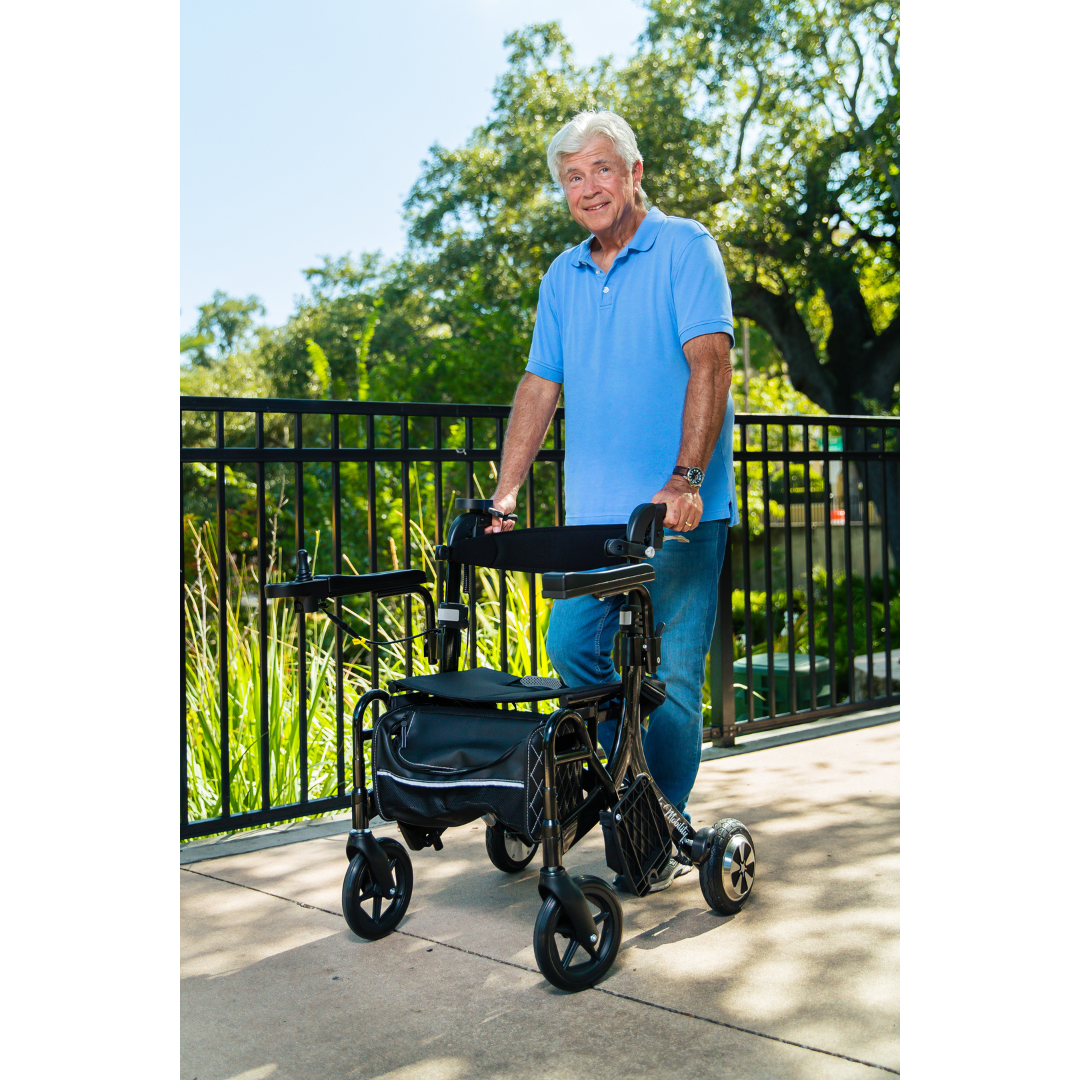 An older man in a blue polo and jeans stands outdoors on a sunny day, smiling as he holds onto a Miracle Mobility Portable 4N1 Walker Wheelchair Combo near a metal fence surrounded by greenery and trees.