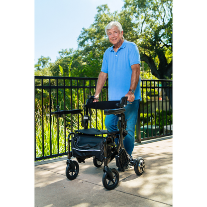An older man in a blue polo and jeans stands outdoors on a sunny day, smiling as he holds onto a Miracle Mobility Portable 4N1 Walker Wheelchair Combo near a metal fence surrounded by greenery and trees.