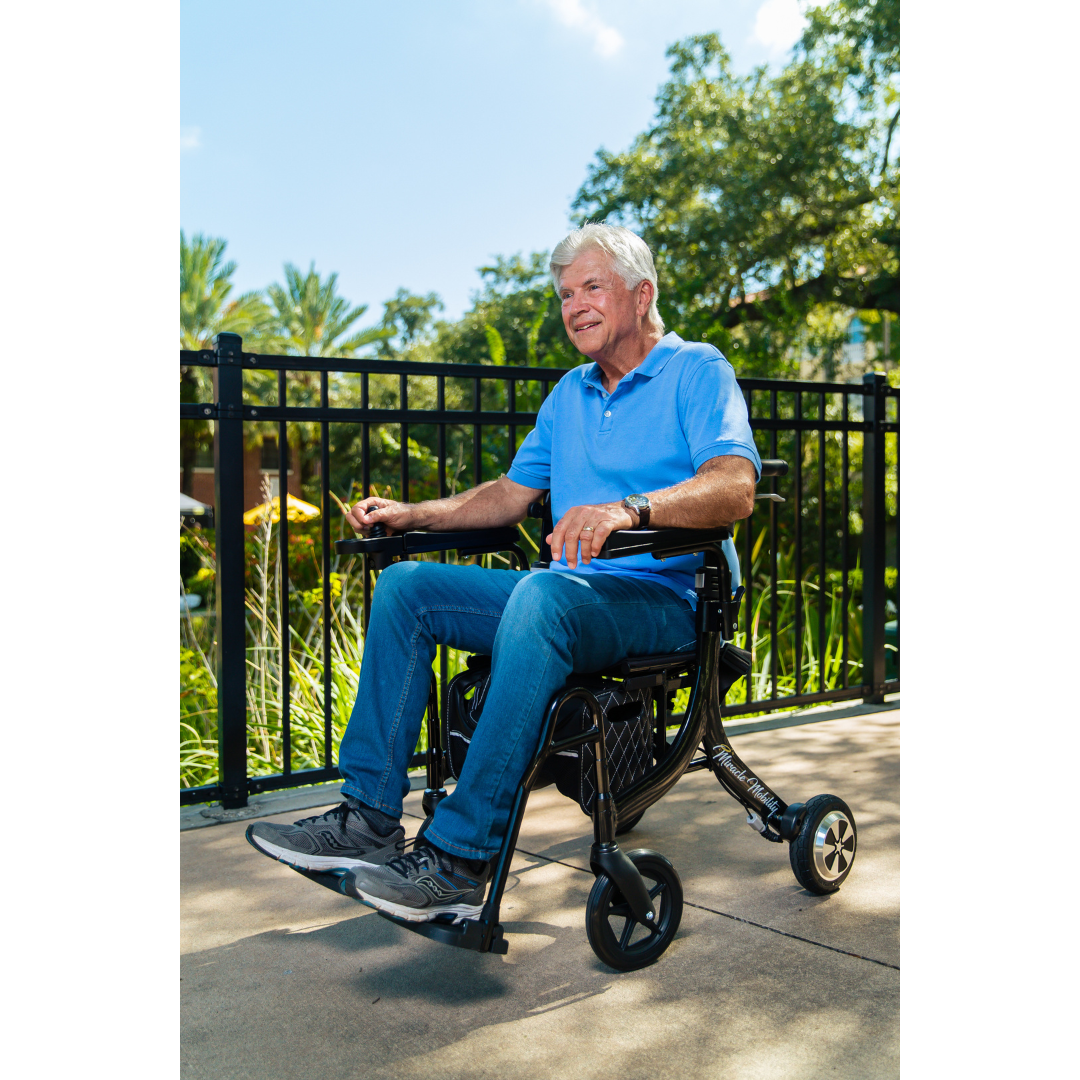 An older man in a blue polo and jeans sits outdoors on a sunny day, smiling in his Miracle Mobility Portable 4N1 Walker Wheelchair Combo, with greenery and a black metal fence behind him.
