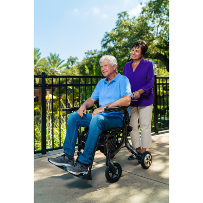 A man in a Miracle Mobility Portable 4N1 Walker Wheelchair Combo smiles as a woman stands behind him, both enjoying a sunny day outdoors by a black fence and green trees.