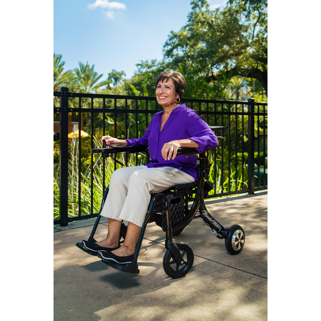 A smiling woman in a purple blouse and beige pants enjoys a sunny day outdoors, sitting in the Miracle Mobility Portable 4N1 Walker Wheelchair Combo with trees and a black metal fence in the background.