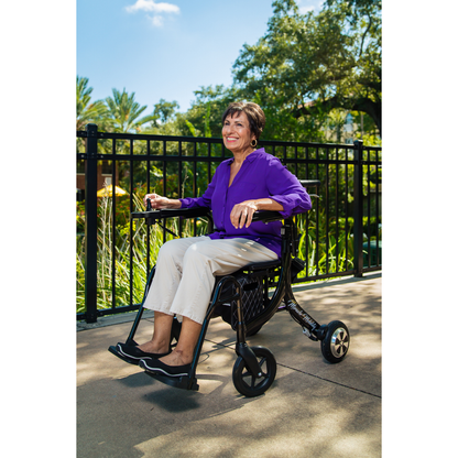 A smiling woman in a purple blouse and beige pants enjoys a sunny day outdoors, sitting in the Miracle Mobility Portable 4N1 Walker Wheelchair Combo with trees and a black metal fence in the background.