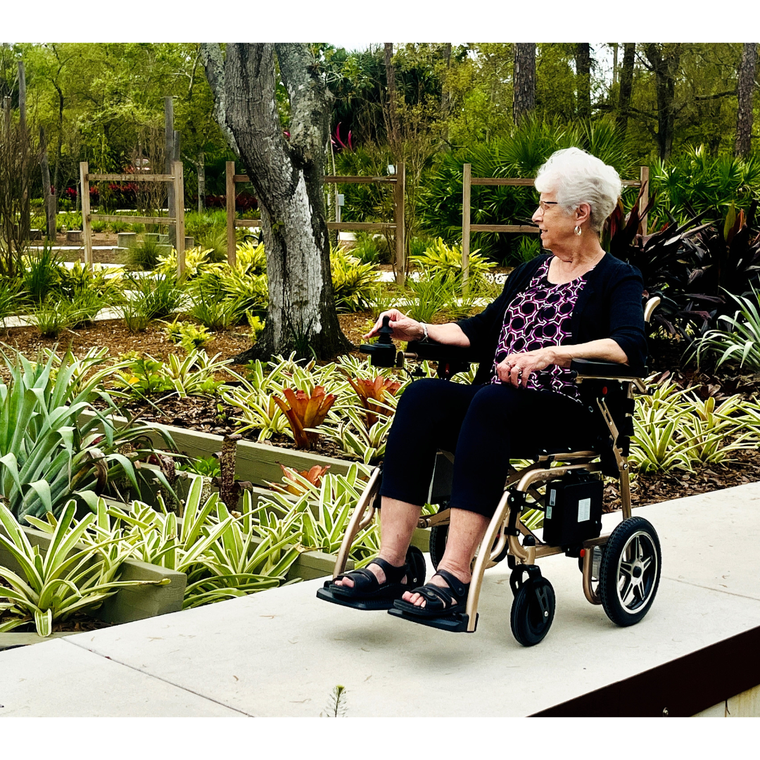 An elderly woman with short white hair sits in a Miracle Mobility Falcon 5000 Lightweight Powerchair on a garden path, surrounded by lush plants and trees, enjoying the peaceful outdoor setting.