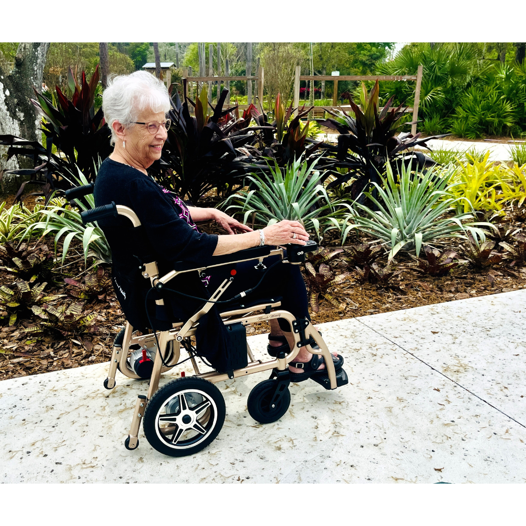 An elderly woman with short white hair smiles while seated in the Miracle Mobility Falcon 5000 Lightweight Powerchair on a paved path, surrounded by lush green and brown plants and trees in a tranquil garden.