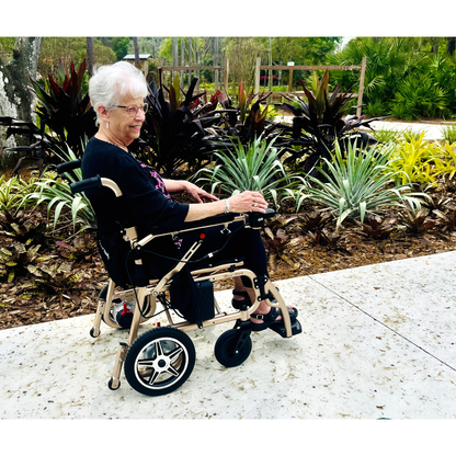 An elderly woman with short white hair smiles while seated in the Miracle Mobility Falcon 5000 Lightweight Powerchair on a paved path, surrounded by lush green and brown plants and trees in a tranquil garden.