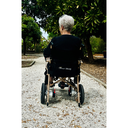 An older adult with short gray hair sits in a Miracle Mobility Falcon 5000 Lightweight Powerchair on a gravel path, surrounded by trees and greenery, seen from behind.