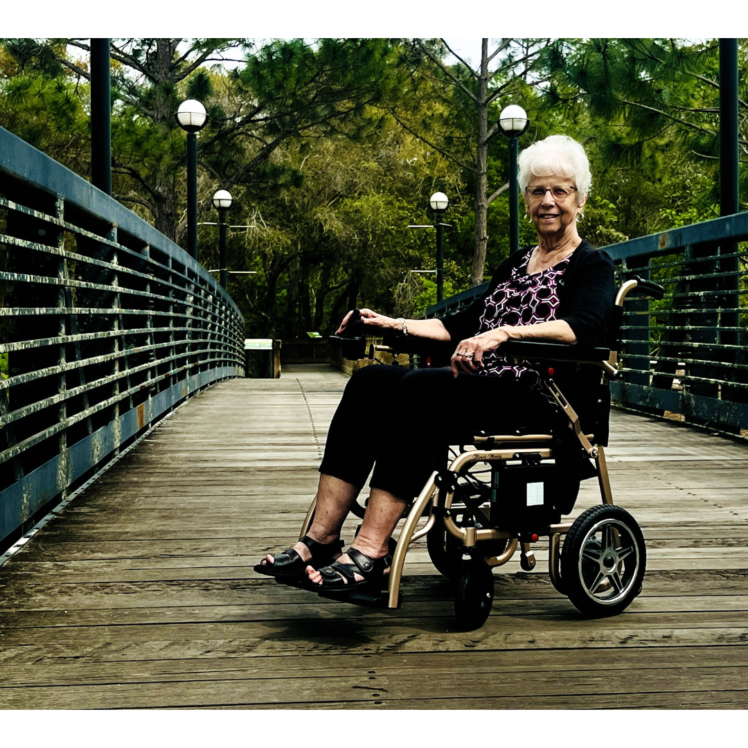 An elderly woman with white hair smiles while seated in a Miracle Mobility Falcon 5000 Lightweight Powerchair on a wooden bridge, surrounded by greenery and trees under round streetlights on a cloudy day.