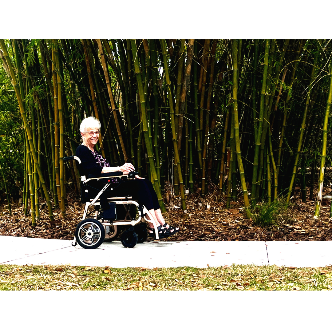 An older woman with short white hair smiles while sitting in a Miracle Mobility Falcon 5000 Lightweight Powerchair on a sidewalk, with tall bamboo plants in the background.