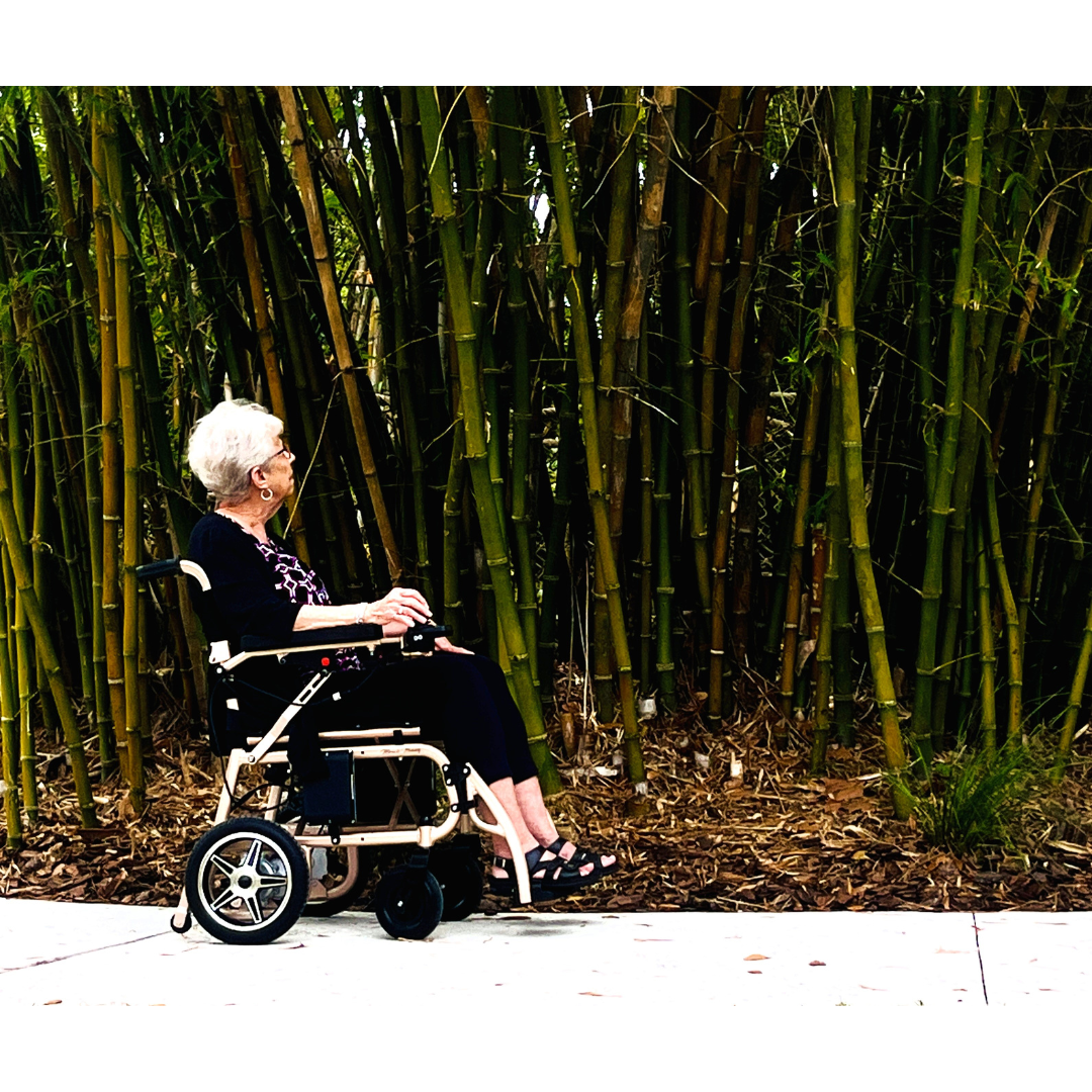 An elderly woman with short white hair sits in a Miracle Mobility Falcon 5000 Lightweight Powerchair on a sidewalk, facing a dense grove of tall bamboo stalks as she enjoys the natural scenery.