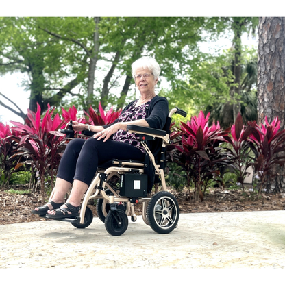 An older woman with white hair smiles while sitting in a Miracle Mobility Falcon 5000 Lightweight Powerchair on a paved path, surrounded by red and green plants and trees in a park setting.