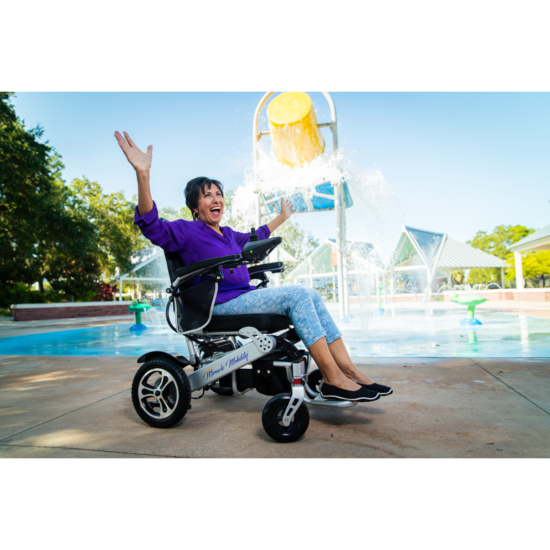 A smiling woman in a purple shirt sits in a Miracle Mobility Palladium 8500 Portable Powerchair with arms raised, enjoying a splash pad fountain on a sunny day at the park.