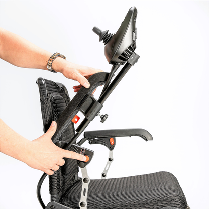 A person adjusts the joystick on the armrest of the Miracle Mobility Palladium 8500 Portable Powerchair, showcasing its foldable design and movable parts with a black mesh finish against a white background.