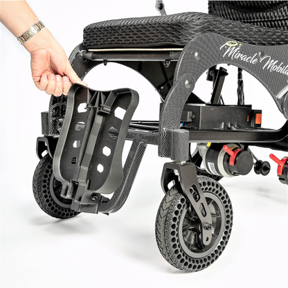 A hand is adjusting or removing the footrest of the Miracle Mobility Palladium 8500 Portable Powerchair, a lightweight, foldable wheelchair with black wheels and a textured frame.
