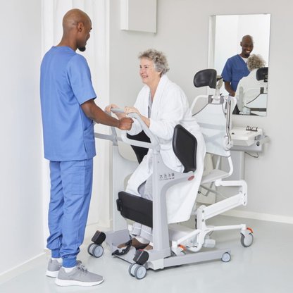 A nurse in blue scrubs assists an older woman in a bathrobe, who is seated on a Molift Transfer Pro – Innovative Sit-To-Stand Patient Lift, in a bright, clean room with a mirror and sink in the background.