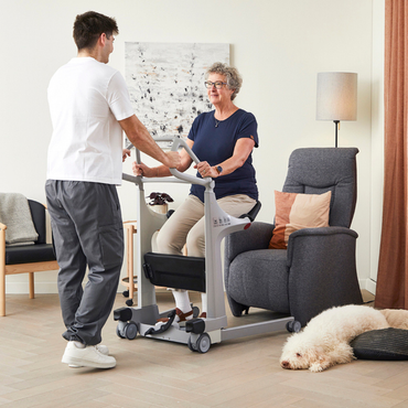 A senior woman uses the Molift Transfer Pro - Innovative Sit-To-Stand Patient Lift, with assistance from a caregiver. She is seated near a gray armchair in a cozy living room, while a fluffy white dog sleeps on the floor nearby.