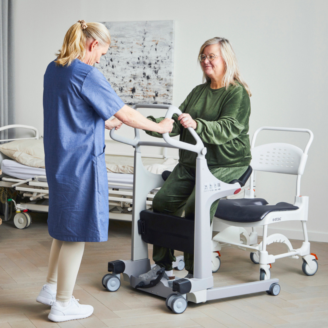 A caregiver assists a seated woman using the Molift Transfer Pro - Innovative Sit-To-Stand Patient Lift in a medical setting, with hospital beds visible in the background.