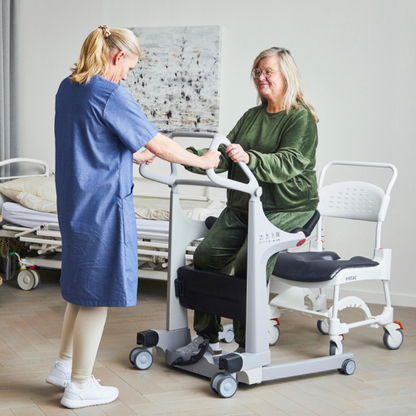 A caregiver assists a seated woman using the Molift Transfer Pro - Innovative Sit-To-Stand Patient Lift in a medical setting, with hospital beds visible in the background.