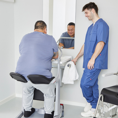 A healthcare worker in blue scrubs assists a seated patient using the Molift Transfer Pro – Innovative Sit-To-Stand Patient Lift in a clinical setting; both appear to be interacting and smiling in front of a mirror.