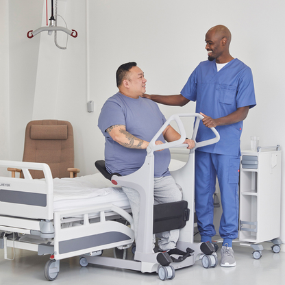 A healthcare worker in blue scrubs assists a patient using the Molift Transfer Pro - Innovative Sit-To-Stand Patient Lift next to a hospital bed, both smiling in a bright, modern medical room.