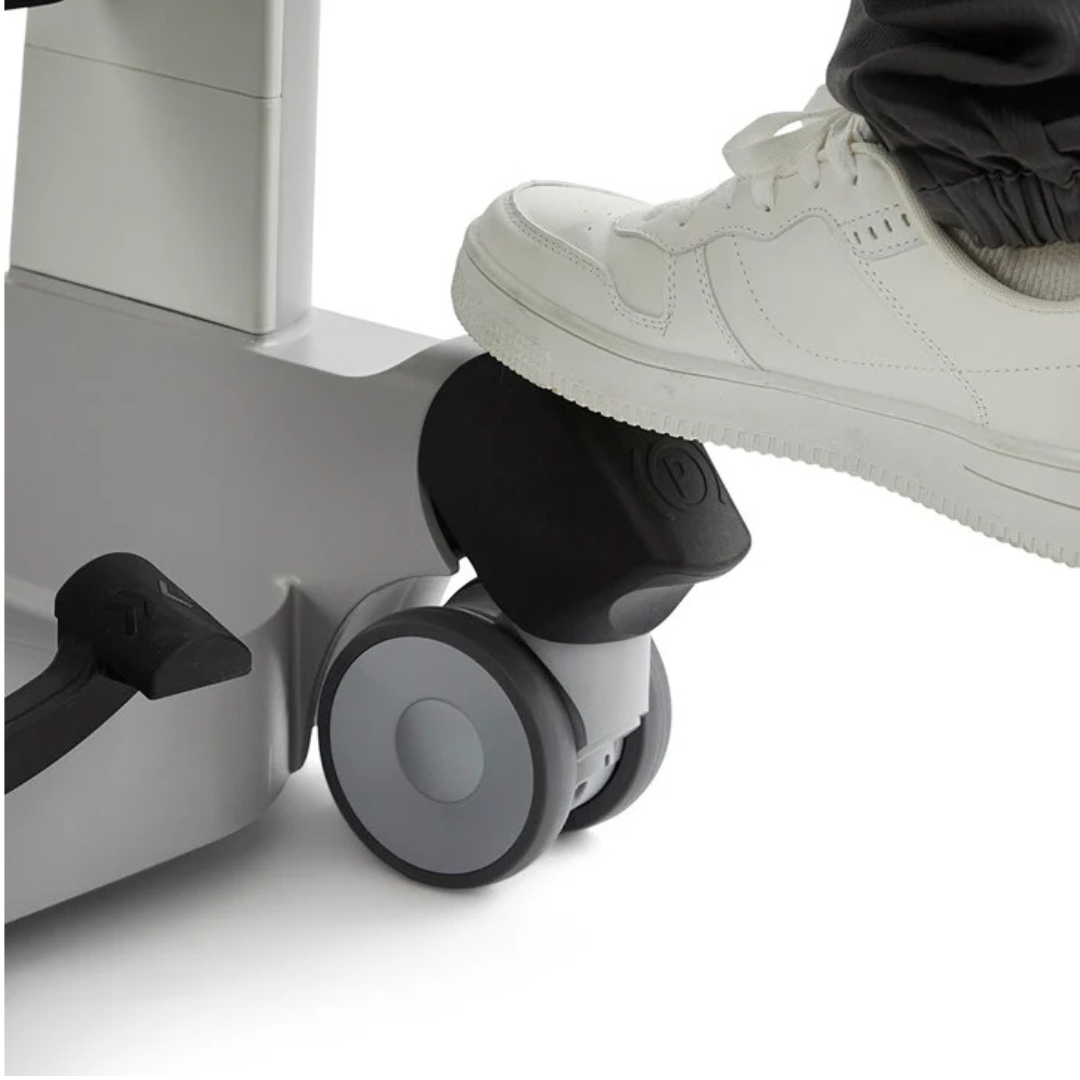 A person’s foot in a white sneaker presses a black pedal attached to the gray wheelbase of a sit-to-stand transfer aid, such as the Molift Transfer Pro - Innovative Sit-To-Stand Patient Lift.