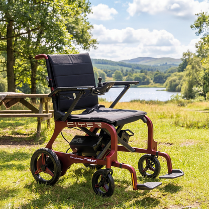 The Monarch Flyer Ultra-Lite Carbon Powerchair, in vibrant red, is shown on grass by a picnic table, set against a beautiful lakeside with trees and hills—an ideal spot to experience its modern features in action.