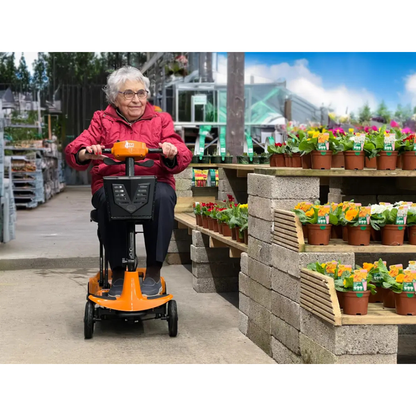 An older woman in a red jacket smiles as she rides the Monarch Genie Ultralight Portable Mobility Scooter through a garden center. However, the product description contains only CSS code and lacks any product details.