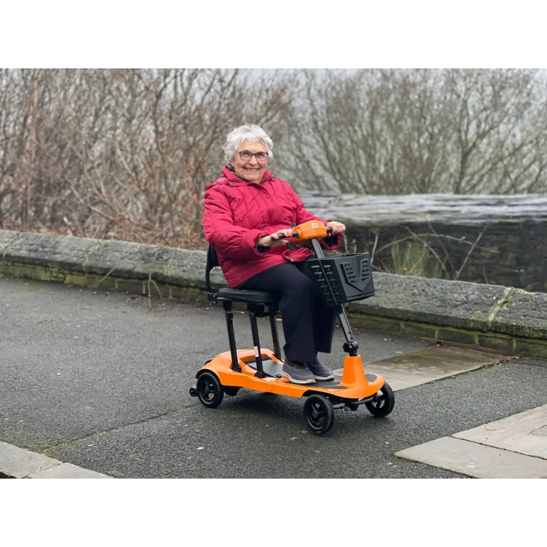 An elderly woman in a red jacket smiles while riding the Monarch Genie Ultralight Portable Mobility Scooter along a paved path, passing bare trees and a stone wall—highlighting her independence and the scooter’s accessibility.